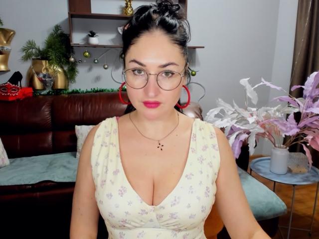 StefanaWinter with glasses, red hoop earrings, black hair in a bun, pink lipstick, yellow floral dress, showing big tits, standing in a room with a brown leather couch.