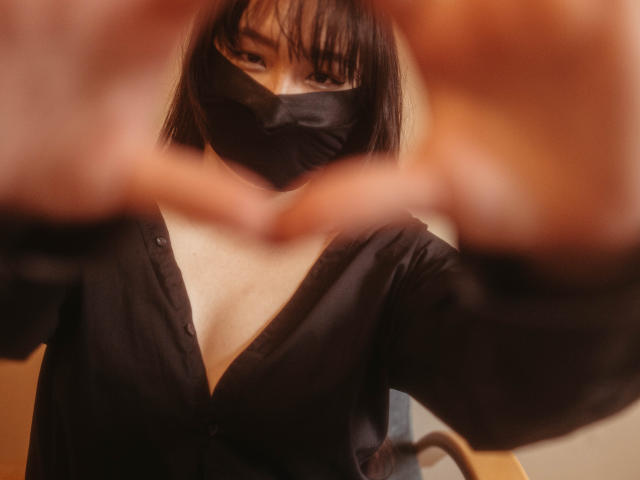BeloverNana with black mask, deep V-neck black shirt, showing cleavage, making heart shape with fingers in foreground, brown eyes, straight black hair, soft lighting, subtle smile.