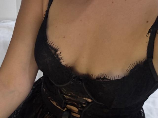 EricaLust's black lace bra with frayed edges, showcasing her tits, has a small black bow in the center and a lace-up detail below her chest.