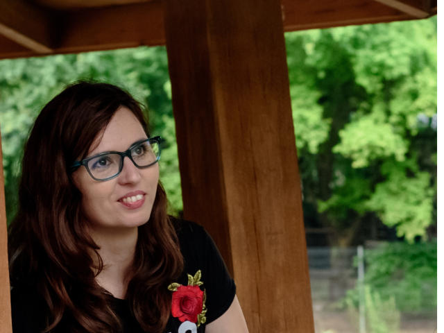 SweetLouise has wavy brown hair, blue glasses, and a black t-shirt with a red rose, smiling under a wooden structure with green trees in the background.