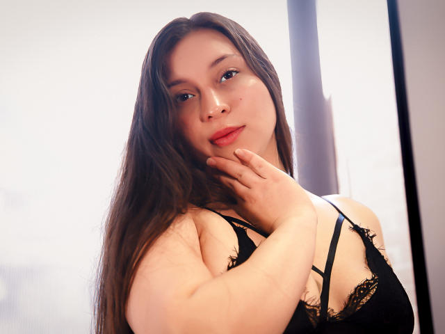 WidnyBeaumont with long brown hair, wearing black lace bra, hand on neck, soft smile, pale skin, red lipstick, looking at camera, light in background, slightly blurry.