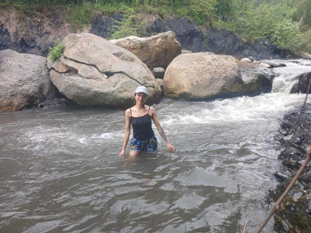 A photo of Kaiix standing waist-deep in a rocky river. Kaiix, a slim woman with short brown hair, wears a white headband, black tank top, and blue floral shorts. She's in the center, with water swirling around her. Large, light brown rocks are in the background, with a small waterfall to the right. The river's surface reflects light, and the greenery above the rocks adds a natural, lush feel. The watermark "ygpix" is in the bottom right corner.