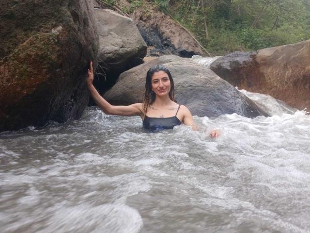 Kaiix, avec ses cheveux bruns mouillés, souriante, portant un maillot de bain noir mouillé, debout dans un ruisseau rocheux, tenant un rocher de la main droite, de l'eau tourbillonnant autour d'elle..