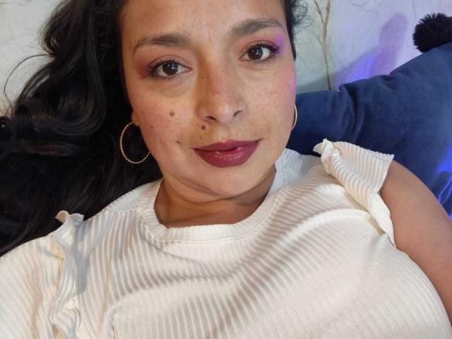 StangelarRivera with dark hair, hoop earrings, pink eyeshadow, red lipstick, white ribbed shirt, and small beauty mark on her right cheek, looking at the camera.