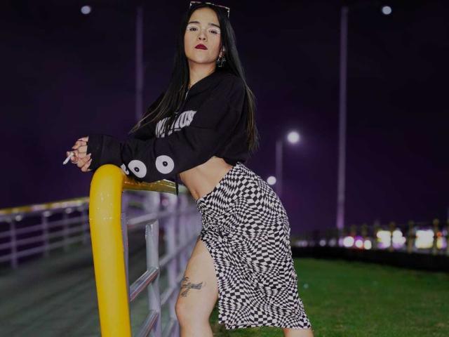 AliceHotBlue with long black hair, wearing a black crop top and houndstooth skirt, leaning on a yellow pole, showing a thigh tattoo, holding a cigarette.