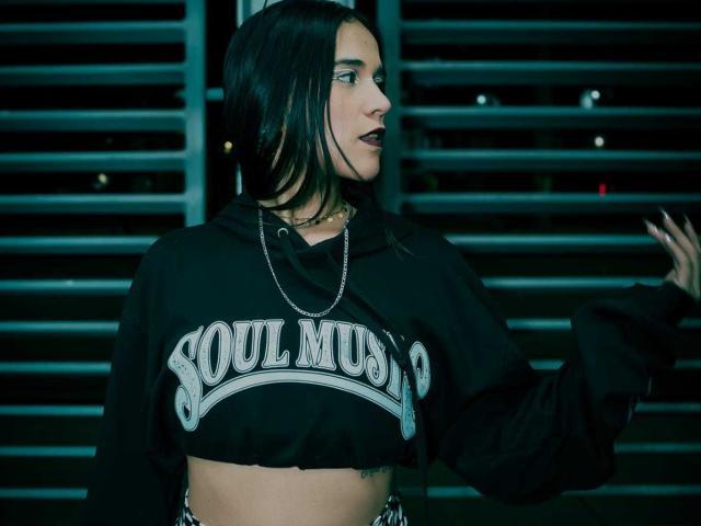 AliceHotBlue with black hair, dark lipstick, wearing a black "SOUL MUSIC" crop hoodie, chain necklace, and a belly button tattoo, in a profile shot, mid-movement.