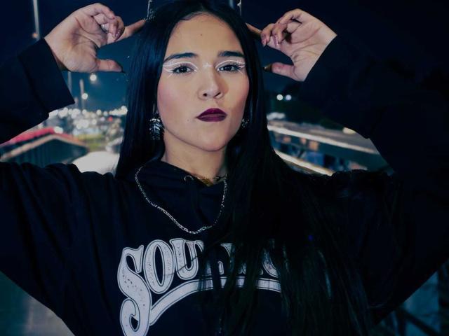 AliceHotBlue with long black hair, dark lipstick, wearing a black hoodie, hands adjusting sunglasses, chain necklace, city lights blurred in the background. She looks confident and stylish.