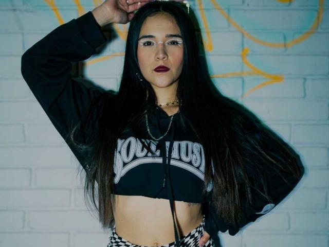 AliceHotBlue with long black hair, wearing a black crop top, showing her tits, black and white checkered pants, hand on head, against a white brick wall.