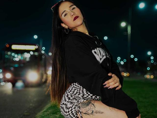 AliceHotBlue with long black hair, red lipstick, black hoodie, checkered skirt, and tattoo on thigh, standing with arms crossed, night, street lights, car lights in background.