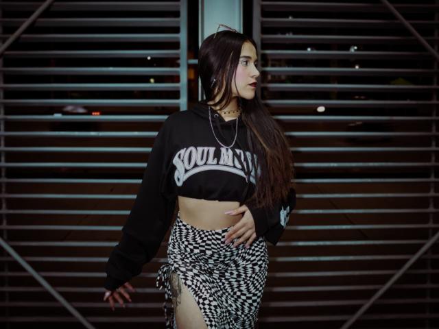 AliceHotBlue with long brown hair, black "Soulboy" crop hoodie, and black-and-white houndstooth skirt, showing her toned midriff and a side slit, standing in a dark urban setting.