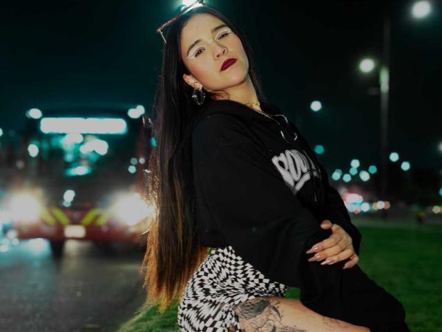 AliceHotBlue with long black hair, wearing a black hoodie and checkered skirt, arms crossed, red lipstick, tattoo on thigh, standing night, city lights, red car in background.