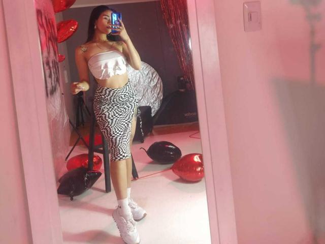AliceHotBlue stands in a mirror, taking a selfie. She's wearing a white bandeau, black and white patterned skirt, and white sneakers. Her ass is small and her tits are medium.