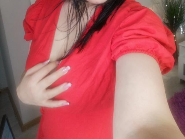 ItsMarry in a red dress with puffy sleeves, holding her left boob with her right hand, has black hair and a fair skin tone. Her nails are painted pink.