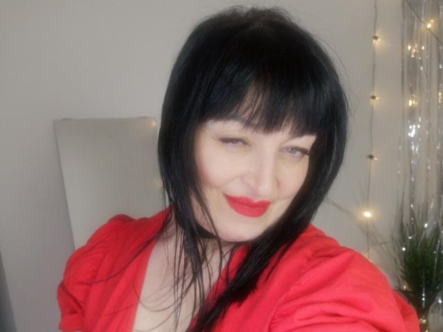 ItsMarry with black hair, red lipstick, and red shirt, smiling slightly, showing cleavage, with a soft expression and subtle makeup, standing in front of a string of lights.