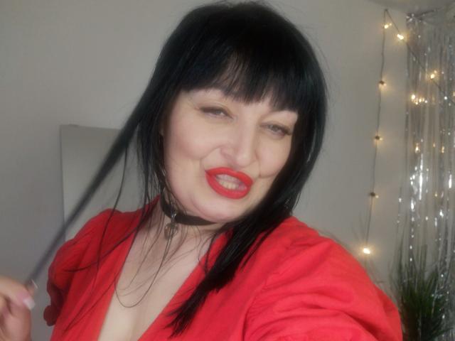 ItsMarry with black hair, red lips, red shirt, and choker, holding a lock of hair, smiling slightly. Boobs visible, pale skin, and a sexy, playful look.