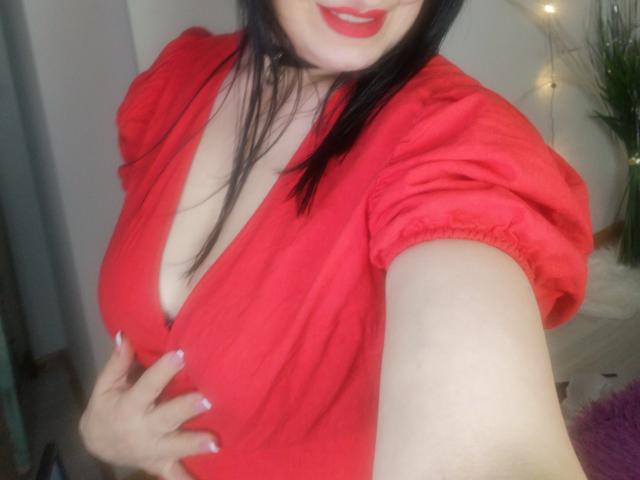 ItsMarry with dark hair, red dress, puffy sleeves, and red lipstick, showing off her big tits, holding her dress down, smiling, with a hand on her chest.