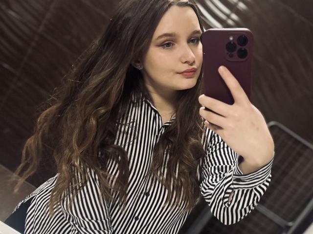 MagikPeachh with wavy brown hair, wearing a striped shirt, taking a selfie with a purple phone, holding it in her right hand, smiling subtly, in a mirror shot.