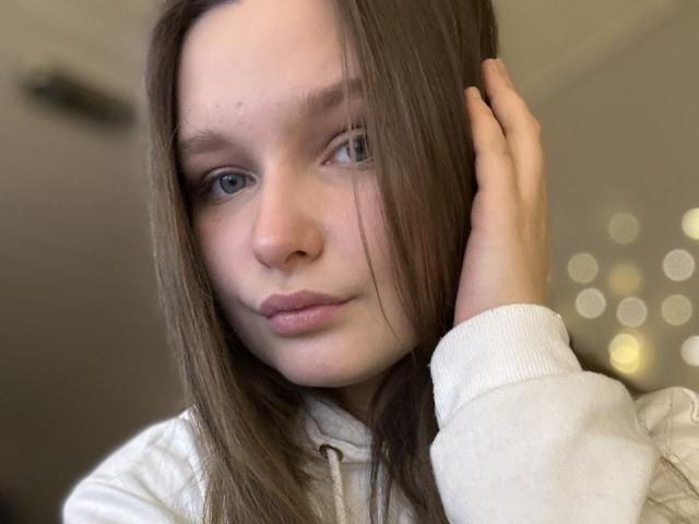 MagikPeachh, close-up portrait with soft lighting. Brown hair, straight and parted to the side, covers one eye. Pale skin, light blue eyes, and slightly pursed lips. Right hand touches hair. Wearing a white, ribbed, long-sleeve hoodie. Blurred, bokeh-style lights in the background. Facial expression is neutral, slightly curious. Image is slightly overexposed, highlighting MagikPeachh's facial features. Simple, natural composition.