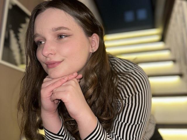 MagikPeachh with long brown hair, striped shirt, hands under chin, smiling gently, light skin, small nose, soft lips, earrings, sitting on stairs, warm lighting, relaxed expression.