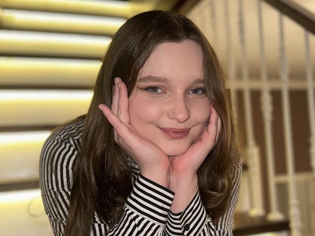 MagikPeachh with hands on cheeks, smiling softly, striped black and white shirt, long brown hair, blue eyes, fair skin, gentle expression, indoors, warm lighting.