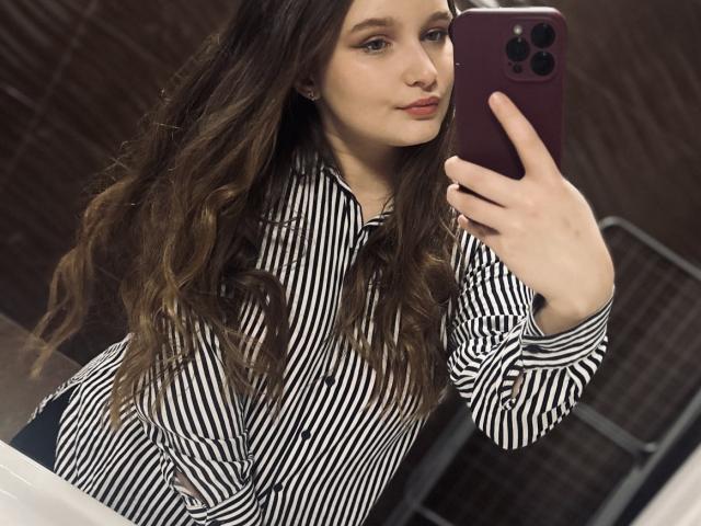 MagikPeachh with wavy brown hair, striped black-and-white shirt, holding purple phone, taking mirror selfie, showing slight pout, fair skin, subtle makeup, and natural tits in bathroom.