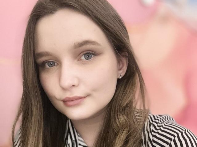 MagikPeachh has fair skin and long brown hair. She has blue eyes, subtle makeup with light eyeshadow, and small pearl earrings. She wears a black-and-white striped shirt. Her expression is neutral with slightly parted lips. The image is a close-up photograph with a soft, pink background blurred in the back. The focus is on MagikPeachh's face. The lighting is even, highlighting her facial features.