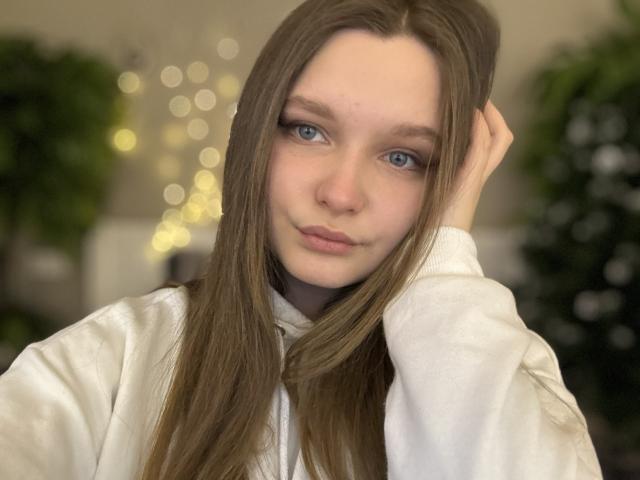 MagikPeachh has long brown hair, blue eyes, and is wearing a white shirt with a serious expression, touching her hair. Boobs are not visible. Soft light, bokeh in background.