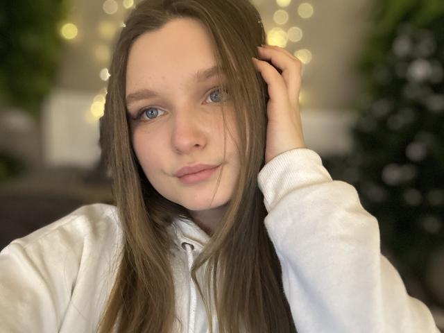 MagikPeachh with long brown hair, blue eyes, and a slight smile, wearing a white hoodie, hand in hair, soft bokeh lights in the background.