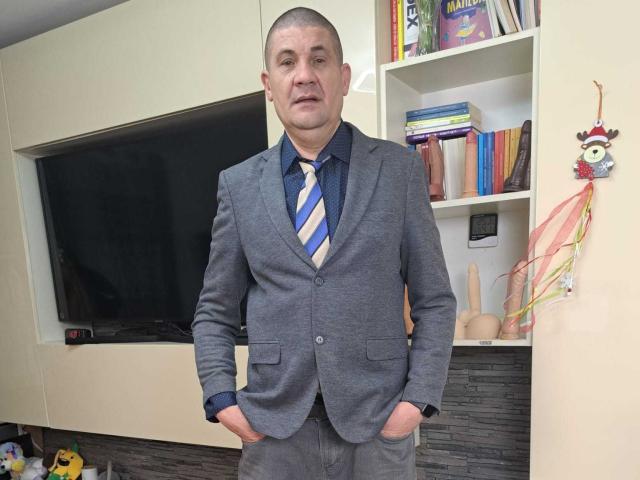 CrisSalon stands in a room, wearing a gray suit with a blue and white striped tie, hands in pockets. He has a bald head, a medium build, and a slightly serious expression. Behind him, a flat-screen TV is mounted on a beige wall, with shelves holding books and a small decorative figure. A colorful ornament hangs on the right. The image has a casual, everyday feel.