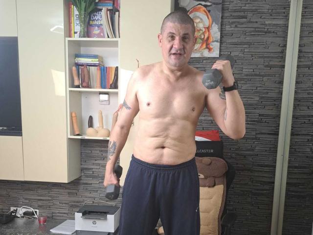 Shirtless CrisSalon with a bald head stands in a room holding a gray dumbbell in each hand. He has a muscular build, visible tattoos on his left arm, and is wearing navy blue pants. The background includes a bookshelf with colorful books and a shelf with sex toys. The wall has a textured gray brick pattern. CrisSalon is looking directly at the camera with a neutral expression. The image is a photograph.