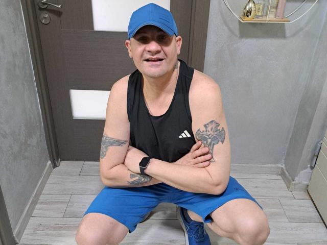 CrisSalon, a muscular man with light skin and short brown hair, squats in a small room. He wears a blue cap, black sleeveless athletic top, blue shorts, and blue sneakers. His arms are crossed over his chest, showing tattoos on both biceps. He has a black smartwatch on his left wrist. The room has a gray door with a white square panel, and a small shelf with a canister is on the right wall. The floor is light wood. CrisSalon's expression is slightly open-mouthed, with a subtle smile.