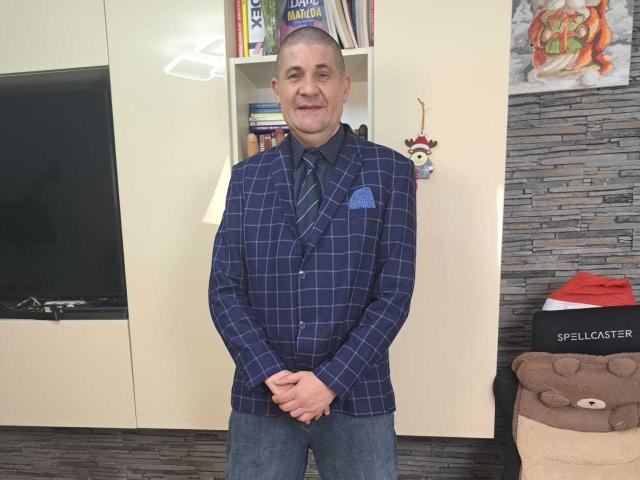 CrisSalon has a bald head, wearing a blue checkered sweater, dark button-up shirt, and jeans, standing with hands clasped, smiling. He's in a cozy room with a TV.