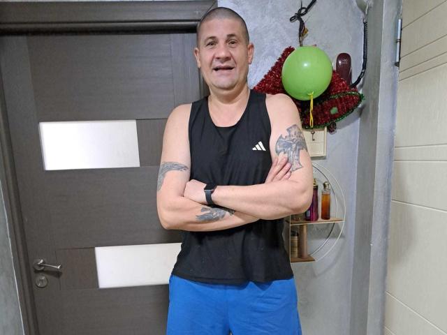CrisSalon, bald with tattoos on both arms, wearing a black sleeveless top and blue shorts, standing with arms crossed, smiling, in front of a wooden door.