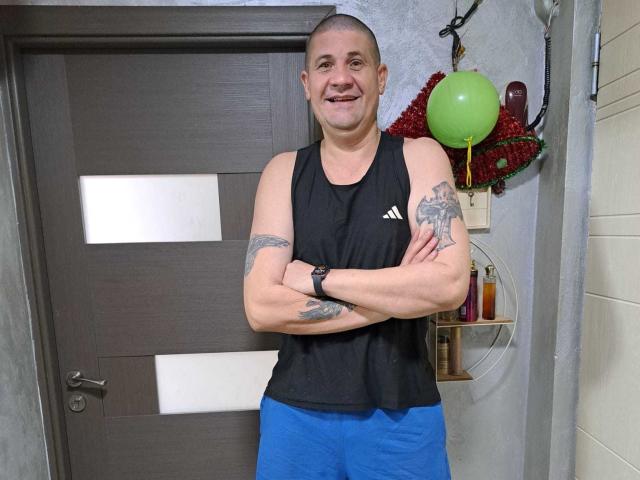 CrisSalon stands in a room, arms crossed, smiling at the camera. He's a bald, muscular man with tattoos on both biceps. He wears a black sleeveless athletic shirt with an Adidas logo and bright blue shorts. His right wrist has a black wristwatch. The background has a wooden door, a green balloon, and red decorations on the wall. The room's walls are textured. CrisSalon's tattoos are detailed, and the overall image is clear and well-lit.
