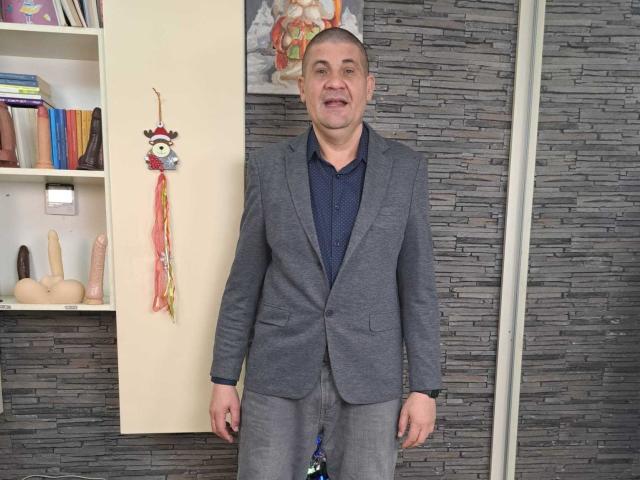 CrisSalon stands in a gray suit, blue shirt, with a visible dildo hanging from his belt. He's middle-aged, smiling, with short gray hair, and a small mustache.
