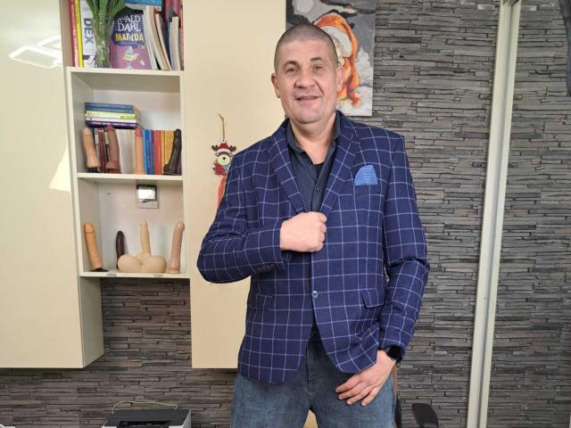 CrisSalon, a middle-aged man with a shaved head, wearing a blue checkered blazer, holds his jacket, smiling, with a blue pocket square, standing in a room.