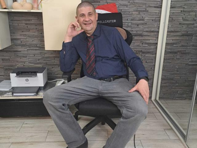 CrisSalon, bald, smiling, wearing blue shirt, red-striped tie, gray pants, sitting with legs spread, hand on knee, holding phone to ear, in office chair, printer on desk.