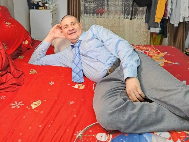 CrisSalon, a middle-aged man with a bald head and a slight smile, lies on a red bed with Christmas-themed patterns. He wears a light blue shirt with a blue checkered tie and gray pants. His right arm is bent, resting on the bed, while his left arm is stretched out. The bed's red cover features holiday motifs. The room includes a white cabinet and a lace curtain in the background. CrisSalon's relaxed posture and casual attire suggest a comfortable, informal setting.