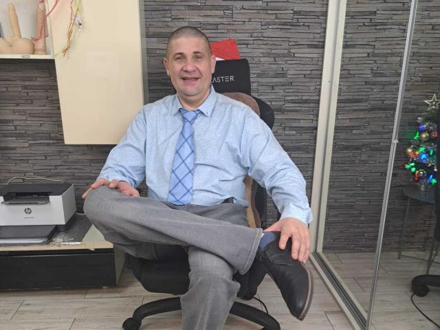 CrisSalon, a middle-aged man with a shaved head and light skin, sits in a black office chair. He wears a light blue patterned shirt with a blue diagonal tie, gray pants, and black shoes. His right leg is crossed over his left. The background includes a gray stone-textured wall, a small Christmas tree with colorful ornaments, and a printer on a shelf. The man smiles slightly, displaying a relaxed posture.