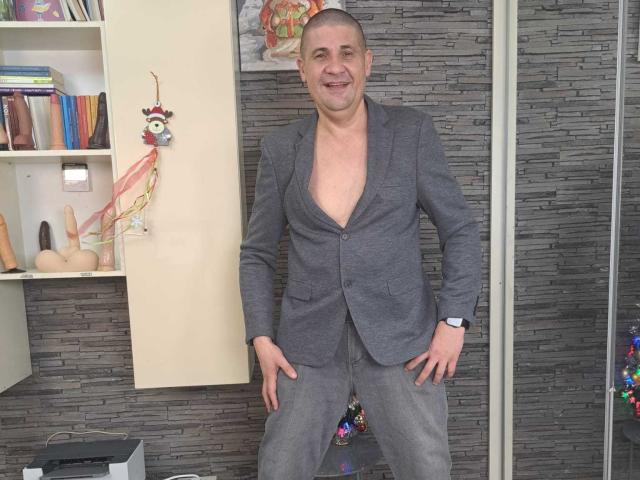 CrisSalon stands in a room, wearing a grey blazer with the bottom button undone, exposing his chest. He has a shaved head, a smiling face, and his hands are on his hips. The blazer is slightly loose, and he wears a watch on his left wrist. The background includes a bookshelf with books, a decorative bird ornament, and a textured grey wall. The room has a playful, slightly cluttered feel.