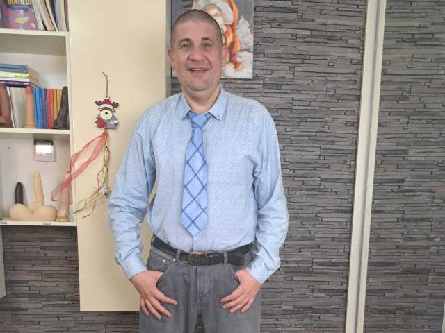 CrisSalon stands in a room, facing the camera with hands on hips. He's a middle-aged man with a shaved head, wearing a light blue, patterned dress shirt and a blue, diamond-patterned tie. He's also wearing gray pants with a black belt. The background features a gray stone-patterned wall and a bookshelf with books, a dildo, and a decorative figure. His shirt is slightly wrinkled, and he's smiling. The image is clear and well-lit.