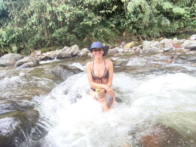 AmelieLautrec, with small tits and a black bikini top, wading in a rocky stream, smiling, holding a yellow drink, wearing a blue hat, with a visible tattoo.