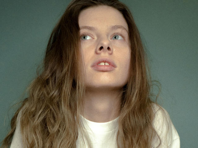 Das Gesicht von Melody Sinclair mit langem, zerzaustem braunen Haar, einer leicht hochgezogenen Nase und leicht geöffneten Lippen, die zur Seite schauen. Sie trägt ein weißes Hemd. Sie hat einen süßen, sanften Ausdruck..