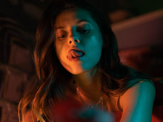 Melody Sinclair mit welligem, braunem Haar, blau-orangefarbener Beleuchtung, leicht herausgesteckter Zunge, die verführerisch blickt, trägt eine Halskette und hat weiche Schatten im Gesicht..