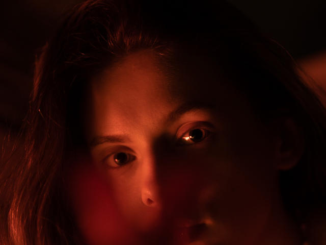Das Gesicht von Melody Sinclair im schwachen, roten Licht, mit weit aufgerissenen, leicht schockierten Augen, und dunklem, braunem Haar, das teilweise beleuchtet ist, wodurch ein stimmungsvoller und intensiver Ausdruck entsteht..