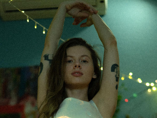 Melody Sinclair met armen omhoog, waarbij tatoeages op haar bovenarmen zichtbaar zijn, draagt een witte top, in een zwak verlichte kamer met lichtjes, haar bruine haar is los, en ze heeft een rustige uitdrukking..