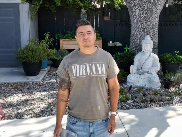 MisterMiggie stands in a backyard, wearing a grey Nirvana t-shirt and blue jeans. He has short, dark hair and a tattoo on his left forearm. A white Buddha statue is to his right, surrounded by small plants and gravel. The background includes a black wooden fence, greenery, and a tree. His relaxed posture and neutral expression dominate the composition. Natural light highlights the scene.