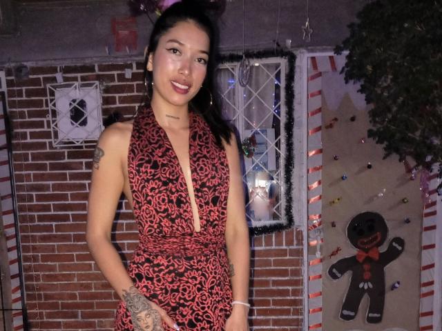 KrystalEvans with black hair, wearing a red and black leopard print dress, showing a tattoo on her right arm, smiling with red lipstick, standing in a festive room.