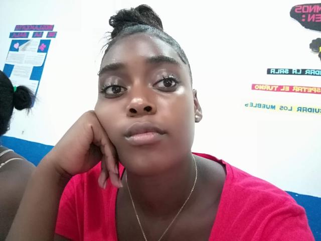 Akemyjhonsson with dark skin, black hair in a bun, wearing a red shirt, silver necklace, and resting head on hand, looking at camera with subtle makeup.