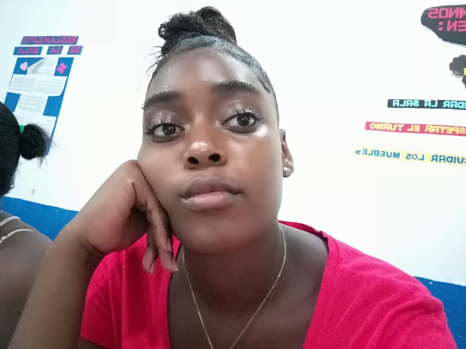 Akemyjhonsson with dark skin, black hair in a bun, wearing a red shirt, silver necklace, and resting head on hand, looking at camera with subtle makeup.